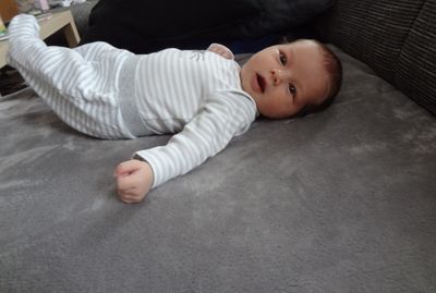 Ein Baby liegt auf einer grauen Decke und trägt ein weiß-grau gestreiftes Langarmshirt. Das Baby schaut mit großen Augen und offenem Mund nach oben.