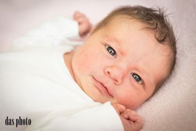 Ein Nahaufnahme eines Neugeborenen, das auf einer rosa Decke liegt, ein weißes Shirt trägt, mit einem leichten Lächeln und blauen Augen, die in die Kamera schauen.