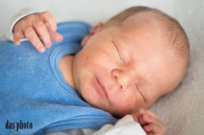 Ein Neugeborenes schläft friedlich auf einer weißen Decke und trägt ein blaues Shirt und ein weißes Langarm-Oberteil. Die Hand des Babys berührt sanft seine Wange.