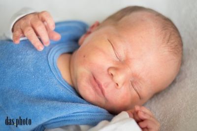Ein Neugeborenes schläft friedlich auf einer weißen Decke und trägt ein blaues Shirt und ein weißes Langarm-Oberteil. Die Hand des Babys berührt sanft seine Wange.