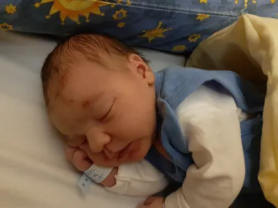 Ein Neugeborenes schläft friedlich auf einem Bett, trägt eine blaue Weste und einen weißen Zettel mit Text.
