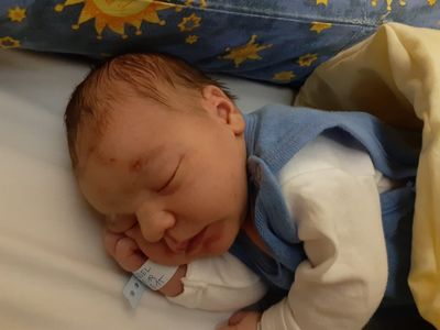 Ein Neugeborenes schläft friedlich auf einem Bett, trägt eine blaue Weste und einen weißen Zettel mit Text.