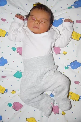 Ein Baby schläft auf einer bunten Decke mit Elefantendesigns, trägt ein weißes Shirt und graue Hosen.