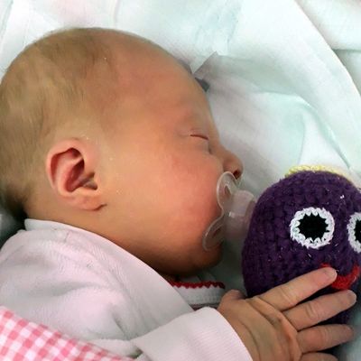 Ein Baby schläft friedlich mit einem Schnuller und einem lila Monster-Plüschtier. Das Baby ist in eine weiße Decke gewickelt.