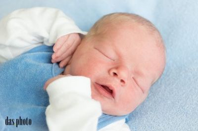 Ein Neugeborenes schläft friedlich auf einer blauen Decke und trägt ein blaues und weißes Outfit. Das Gesicht des Babys ist leicht geöffnet, und es hat eine kleine Hand auf der Wange.