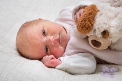 Ein Babymädchen liegt auf einer weißen Decke und hält ein Plüschhundespielzeug. Sie schaut direkt in die Kamera mit einem neugierigen Ausdruck.