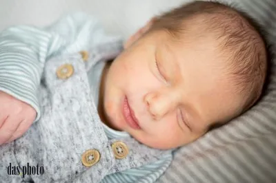 Ein Babyjunge schläft friedlich auf einem Bett und trägt ein blau-weiß gestreiftes Outfit mit goldenen Knöpfen.