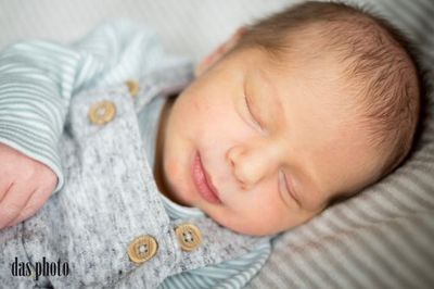 Ein Babyjunge schläft friedlich auf einem Bett und trägt ein blau-weiß gestreiftes Outfit mit goldenen Knöpfen.
