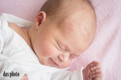Ein Neugeborenes schläft friedlich auf einer rosa Decke und trägt ein weißes T-Shirt. Die Augen des Babys sind geschlossen, und es hat ein leichtes Lächeln auf dem Gesicht.