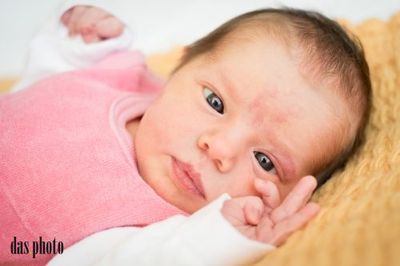 Ein neugeborenes Babymädchen mit rosa Kleidung und blauen Augen liegt auf einer gelben Decke und schaut neugierig in die Kamera.
