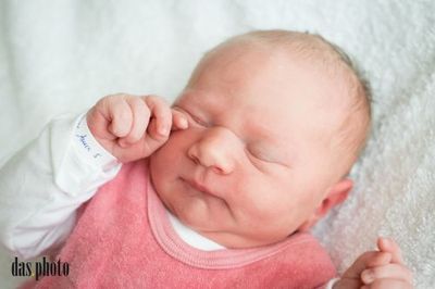 Ein neugeborenes Babymädchen schläft friedlich auf einer weißen Decke und trägt ein rosa Shirt und ein weißes Langarmshirt. Sie hat die Hand auf dem Gesicht.