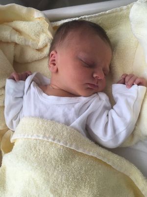 Ein Baby schläft friedlich auf einer gelben Decke, trägt ein weißes Langarmshirt und eine Halskette.