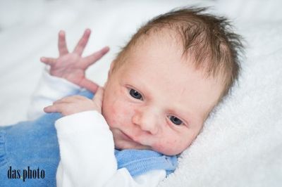 Ein Nahaufnahme eines Babys, das auf einer weißen Decke liegt, ein blaues Outfit trägt, mit einer erhobenen Hand und der anderen auf der Wange, lächelnd.