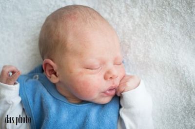 Ein Neugeborenes liegt auf einer weißen Decke und schläft friedlich mit einer Hand in der Nähe des Mundes. Das Baby trägt ein blaues Lätzchen und weiße lange Ärmel.