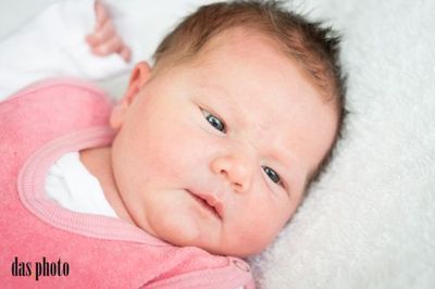 Ein Nahaufnahme eines Babymädchens, das auf einer weißen Decke liegt, ein rosa Pullover trägt, mit leicht geöffnetem Mund und blauen Augen, die direkt in die Kamera schauen.