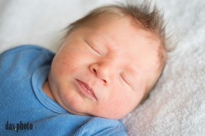 Ein Neugeborenes schläft friedlich auf einer weißen Decke und trägt ein blaues Shirt. Das Gesicht des Babys ist leicht verschwommen, aber sein friedliches Aussehen ist erkennbar.