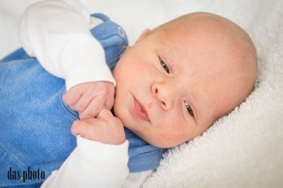 Ein Neugeborenes liegt auf einer weißen Decke, trägt ein blaues Outfit und hat eine Hand auf der Wange. Das Gesicht des Babys ist leicht nach rechts gedreht und scheint etwas anzuschauen.