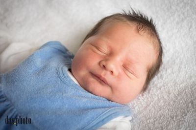 Ein Baby schläft friedlich auf einer weißen Decke und trägt einen blauen Strampler, mit leicht geöffnetem Mund und geschlossenen Augen.