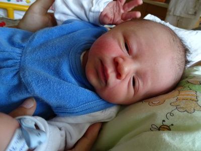 Ein Baby in Blau liegt auf einem Bett, lächelt und schaut in die Kamera, während es von jemandem in einem weißen Hemd gehalten wird.