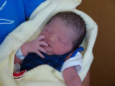 Ein Neugeborenes, eingewickelt in ein gelbes Handtuch, trägt ein blaues Shirt und hält einen Schnuller. Das Baby schläft mit der Hand im Gesicht.