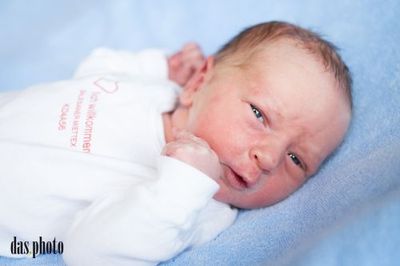 Ein Neugeborenes liegt auf einer blauen Decke und trägt ein weißes Shirt mit rotem Text. Das Baby hat leicht geöffneten Mund und schaut nach rechts.