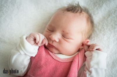 Ein Nahaufnahme eines schlafenden Babymädchens mit einem rosa Shirt und weißen Ärmeln, auf einer weißen Decke liegend.