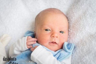 Ein Neugeborenes liegt auf einer weißen Decke, trägt ein blaues Outfit und schaut neugierig in die Kamera.
