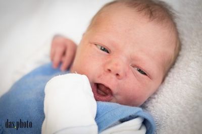 Ein Nahaufnahme eines Neugeborenen, das auf einer weißen Decke liegt, ein blaues Outfit trägt und mit leicht geöffnetem Mund einen überraschten Ausdruck zeigt.