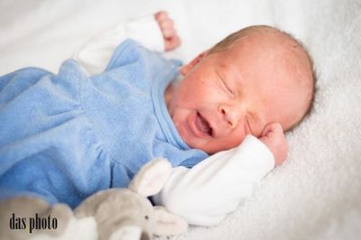 Ein Neugeborenes in einem blauen Outfit liegt auf einer weißen Decke, lächelt und hält ein Kuscheltier.