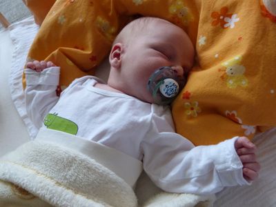 Ein schlafendes Baby mit einem Schnuller im Mund, auf einer gelben Decke mit Blumenmuster liegend.