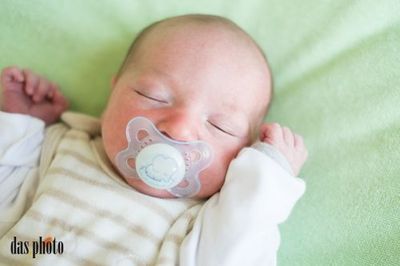 Ein Baby mit Schnuller schläft auf einer grünen Decke. Das Baby trägt ein weiß-beiges gestreiftes Outfit.