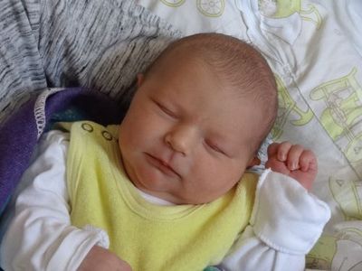 Ein schlafendes Baby in einem gelben Lätzchen und einem weißen Langarmshirt liegt auf einem Bett mit einer gemusterten Decke.