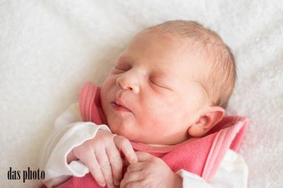 Ein neugeborenes Mädchen schläft friedlich auf einer weißen Decke und trägt eine rosa Jacke und ein weißes Langarmshirt. Sie hat ihre Hände sanft ineinander verschränkt.