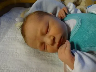 Ein Neugeborenes schläft friedlich auf einem Bett, trägt ein grünes Lätzchen und ein weißes Langarmshirt. Die Hand des Babys liegt auf seinem Gesicht.