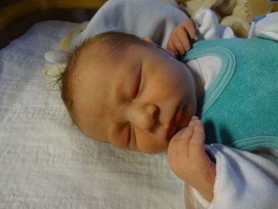 Ein Neugeborenes schläft friedlich auf einem Bett, trägt ein grünes Lätzchen und ein weißes Langarmshirt. Die Hand des Babys liegt auf seinem Gesicht.