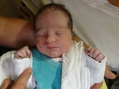 Ein Neugeborenes wird in einem Krankenhausbett gehalten, trägt ein blaues Shirt und eine weiße Decke. Die Augen des Babys sind geschlossen.