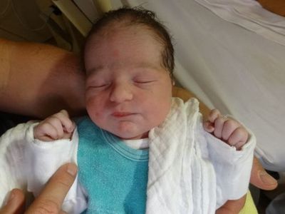 Ein Neugeborenes wird in einem Krankenhausbett gehalten, trägt ein blaues Shirt und eine weiße Decke. Die Augen des Babys sind geschlossen.