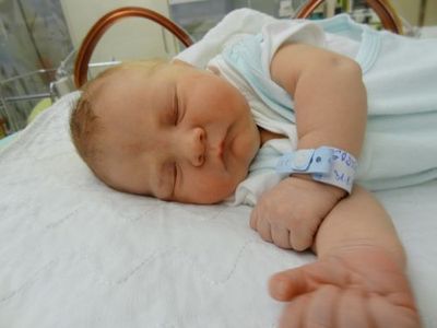 Ein schlafendes Neugeborenes liegt auf einem Krankenhausbett und trägt ein blaues Krankenhausarmband. Das Baby ist in hellblauer und weißer Kleidung gekleidet.