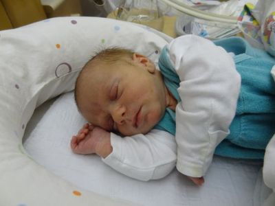 Ein Neugeborenes schläft friedlich in einem Krankenhausbett und trägt ein blau-weißes Outfit. Das Baby ist von einem weißen Kissen mit bunten Punkten umgeben.