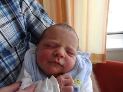 Ein Neugeborenes wird von einem Mann in einem blau-weiß karierten Hemd gehalten. Das Baby hat eine blaue Schürze mit einem gelben Etikett.