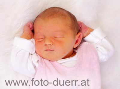 Ein Neugeborenes schläft friedlich auf einer weißen Pelzdecke, trägt eine rosa karierte Schürze und weiße lange Ärmel. Die Hände des Babys liegen sanft auf seinen Ohren.