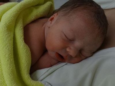 Ein Neugeborenes schläft auf einer grünen Decke, die um seinen Körper gewickelt ist. Die Augen des Babys sind geschlossen, und es scheint in einem friedlichen Schlaf zu sein.