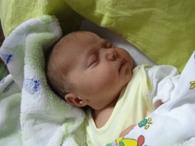 Ein Baby schläft friedlich auf einem Bett, das mit einer grün-weißen Decke bedeckt ist. Das Baby trägt ein gelbes T-Shirt mit einem Cartoon-Charakter darauf.