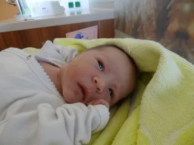 Ein Baby in einem weißen Outfit liegt auf einer gelben Decke und schaut mit einem leichten Lächeln zur Kamera. Hinter dem Baby befindet sich eine Holztheke mit einer rosa Karte.