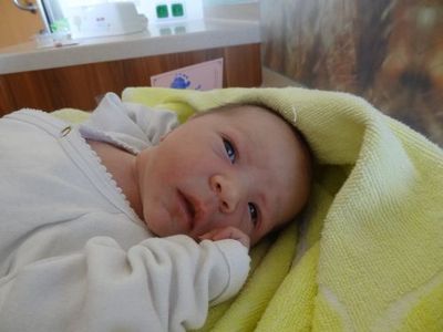 Ein Baby in einem weißen Outfit liegt auf einer gelben Decke und schaut mit einem leichten Lächeln zur Kamera. Hinter dem Baby befindet sich eine Holztheke mit einer rosa Karte.