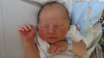 Ein Neugeborenes schläft auf einem Bett mit einer blauen Decke und einem weißen Kissen. Das Baby trägt ein weißes Shirt und eine blaue Armband.