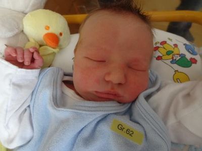 Ein Neugeborenes schläft friedlich in einem Kinderbett, trägt ein blaues Strampler mit einem gelben Etikett 'Gr. 62'. Ein gelbes Plüschtier ist in der Nähe.