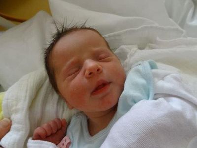 Ein Neugeborenes schläft friedlich auf einer weißen Decke und trägt ein hellblaues T-Shirt. Die Augen des Babys sind geschlossen, und es hat ein leichtes Lächeln im Gesicht.