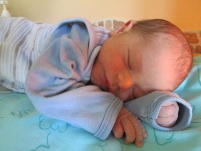 Ein schlafendes Baby in einem blau-weißen Outfit liegt auf einer grünen Decke mit einem Blumenmuster.
