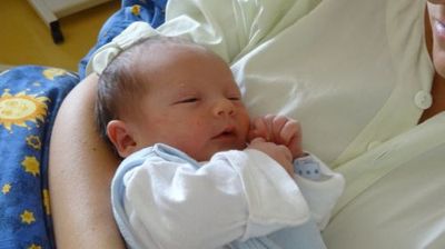 Eine Frau hält ein schlafendes Neugeborenes in ihren Armen, beide tragen helle Kleidung. Das Baby hat die Hände in der Nähe des Gesichts, und die Frau lächelt.